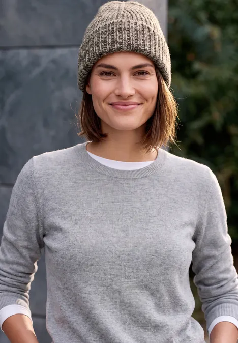 Junge Frau mit grauem Pullover und Strickmütze lächelnd vor unscharfem Hintergrund aus Stein und Grün.