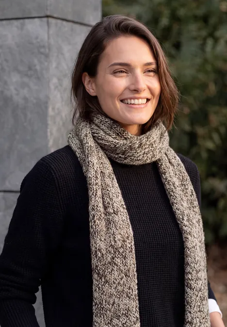 Mouliné Schal truffle brown Junge Frau mit Schal und schwarzem Pullover lächelt im Freien vor einer Steinmauer.