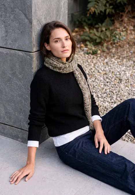 Mouliné Schal truffle brown Frau in schwarzem Pullover, Jeans und Schal lehnt an Mauer, sitzt auf Kiesboden