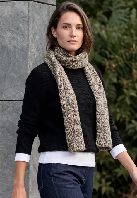 Mouliné Schal truffle brown Frau in schwarzem Pullover, Jeans und beiger Strickschal posiert vor einer Natur- und Steinmauerkulisse.