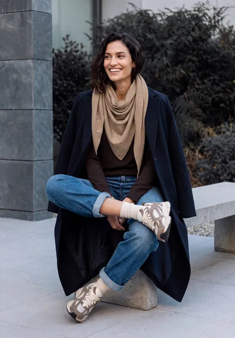 Dreieckstuch mit Glitzerdetails creamy mocha melange Eine lächelnde Frau sitzt im Freien gekreuzt auf einer Bank, trägt Mantel, Jeans, Schal und Sneaker.