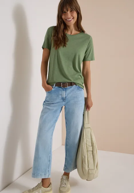 Ceinture Basic à boucle carrée Cacao Frau lehnt an Wand, trägt grünes T-Shirt, weite blaue Jeans mit Gürtel und hält lässige Tasche.