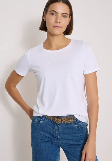 Eine Frau mit kurzen braunen Haaren trägt ein weißes T-Shirt, blaue Jeans und einen Leo-Gürtel.