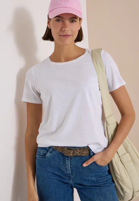 Frau in weißem T-Shirt, Jeans und rosa Cap mit Umhängetasche, stehend vor heller Wand.
