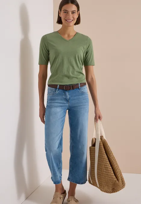 Ceinture à rayures Suede Brown Frau im grünen V-Shirt, hellblauen Jeans, braunem Gürtel und großer Korbtasche.