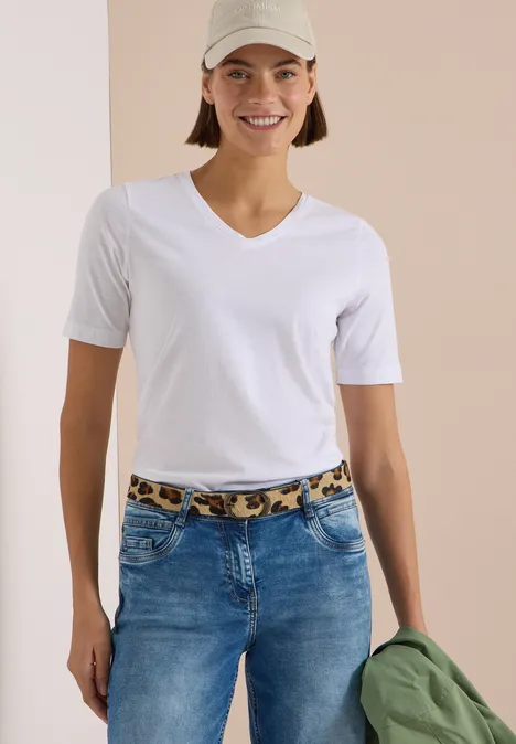 Ceinture en fausse fourrure à motif léopard Natural Leo Print Leather Frau in weißem V-Ausschnitt-T-Shirt und Jeans mit Leoparden-Gürtel, trägt Cap und hält grüne Jacke.