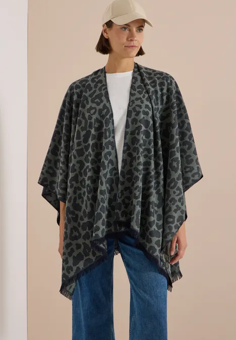 Frau mit gemustertem Poncho-Cardigan, weißem T-Shirt, Jeans und Baseballcap vor hellem Hintergrund.
