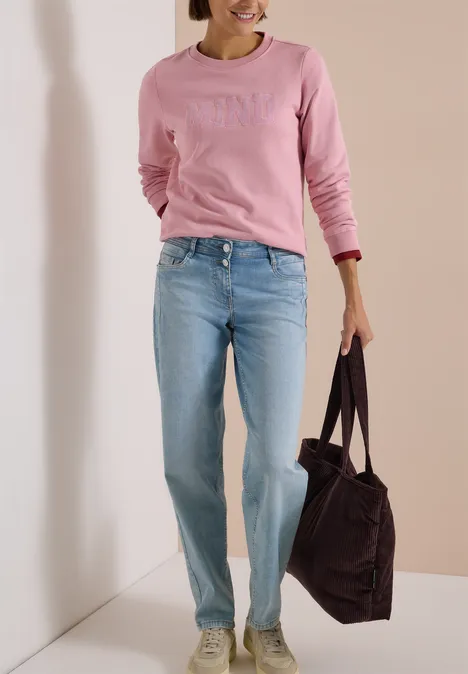 Frau in rosa Sweatshirt und hellblauen Jeans trägt eine große dunkelbraune Tasche vor neutralem Hintergrund.