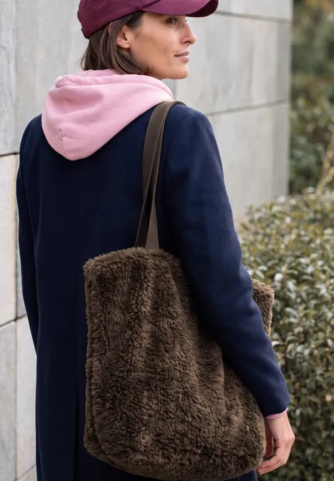 Sac Shopper chocolate sauce brown Femme vue de dos, vêtue d’un manteau bleu marine, d’un sweat à capuche rose et d’un grand sac marron duveteux.