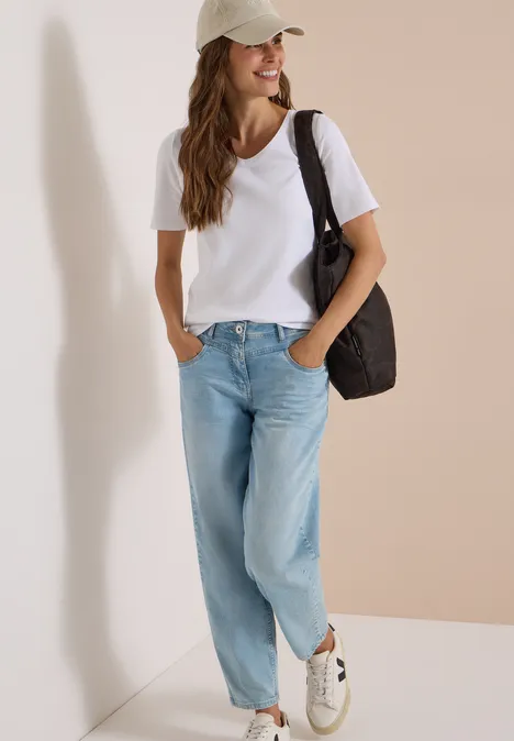 Frau in weißem T-Shirt, hellen Jeans, Sneakers und Baseballcap mit Schultertasche vor neutralem Hintergrund.