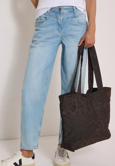 Person in hellblauen Jeans und weißen Sneakern hält eine dunkel gemusterte Stofftasche.