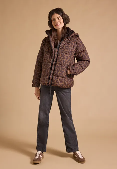 Kuschelige Teddy-Ohrwärmer chocolate sauce brown Frau mit lockigem Haar in brauner gemusterter Steppjacke, dunklen Jeans und weißen Schuhen vor beigem Hintergrund.