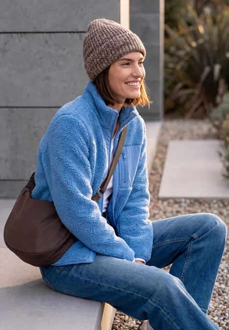 Sac à bandoulière berry red Une femme sourit assise dehors, vêtue d'une veste polaire bleue, jean, bonnet et sac en bandoulière.