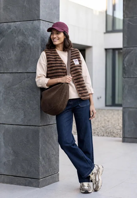 Sac à bandoulière chocolate sauce brown Jeune femme, casquette rouge, doudoune et sac en bandoulière, appuyée contre un pilier gris, souriante.