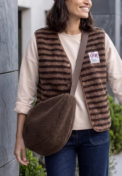Sac à bandoulière chocolate sauce brown Femme portant un gilet rayé marron, un t-shirt clair, un jean et un sac en peluche marron, souriante.