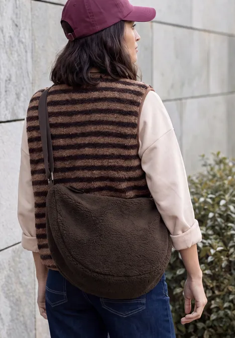 Sac à bandoulière chocolate sauce brown Femme de dos portant un gilet rayé, jean, casquette bordeaux et grand sac en polaire marron.