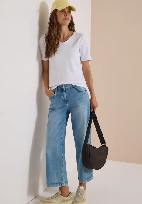 Eine Frau in weißem T-Shirt, weiten Jeans und gelber Mütze lehnt lässig an einer Wand und hält eine Tasche.