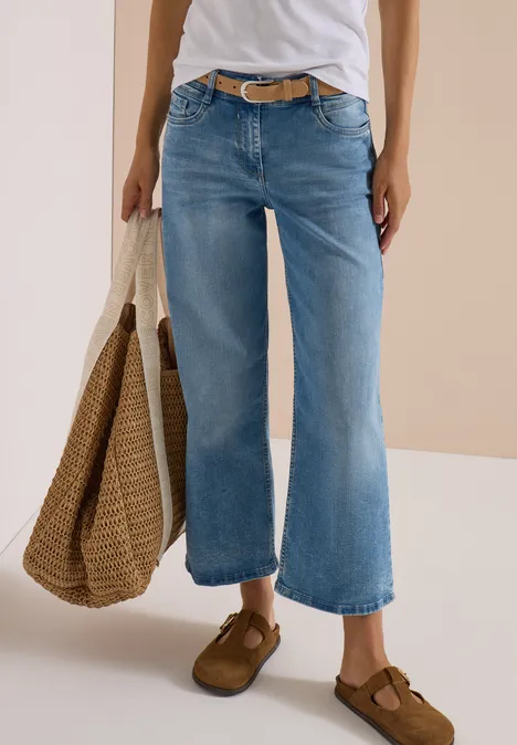 Person trägt weißes Shirt, weite hellblaue Jeans, braune Schuhe und hält eine Strohtasche.