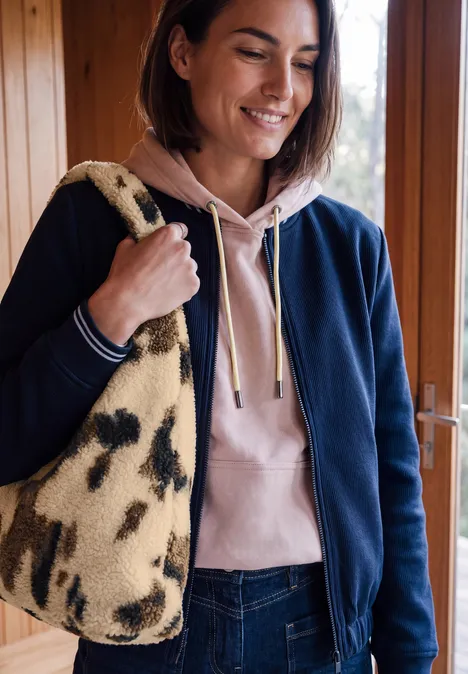 Sac en peluche avec motif de vache pearl beige Une femme souriante en sweat à capuche et veste, portant un sac beige à motifs noirs sur l’épaule.