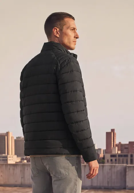 Ein Mann in schwarzer Steppjacke steht auf einem Dach und blickt in die Ferne vor urbaner Skyline.