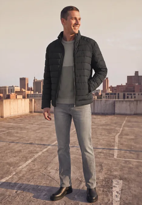 Ein Mann in schwarzer Steppjacke, grauem Hoodie und Jeans steht auf einem Dachparkplatz mit Stadtkulisse.