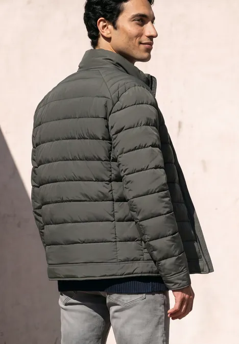 Ein Mann trägt eine graue Steppjacke, steht seitlich und blickt über die Schulter auf hellem Hintergrund.