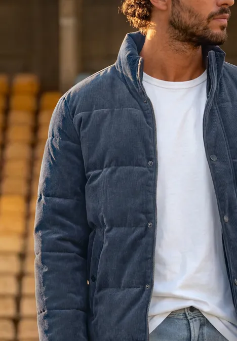 Ein Mann trägt eine dunkelblaue Cord-Steppjacke über weißem T-Shirt, stehend vor Stufen.