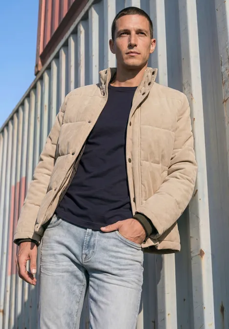 Ein ernst blickender Mann in beiger Pufferjacke und Jeans lehnt an Containern.