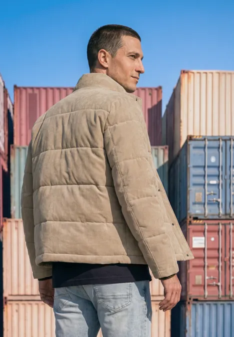 Ein Mann in beiger Steppjacke steht vor bunten Containerwänden, Blick seitlich, urbane Szenerie.
