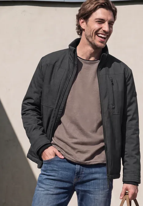 Un homme souriant en veste noire, t-shirt gris et jean bleu.