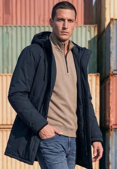 Un homme en parka sombre et jean pose devant des conteneurs colorés empilés.