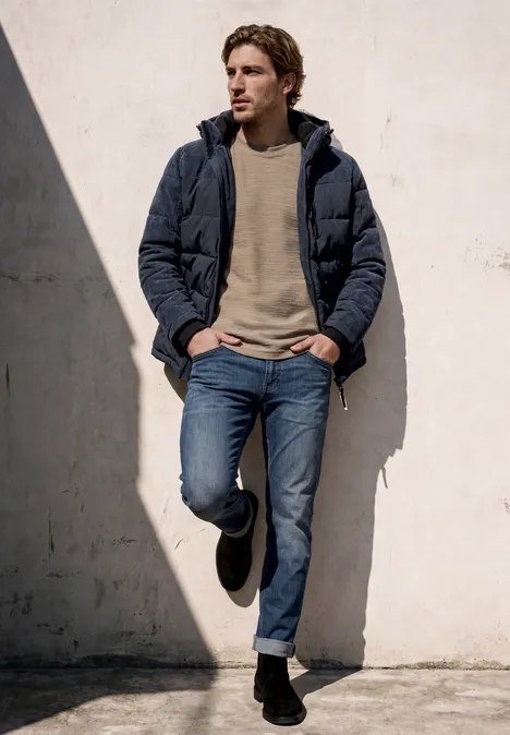 Ein junger Mann lehnt lässig in Jeans und Steppjacke an einer sonnigen Wand.