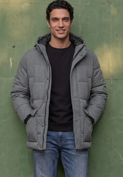 Ein Mann steht vor einer grünen Wand, trägt eine graue Steppjacke mit Kapuze, schwarzen Pullover und Jeans.
