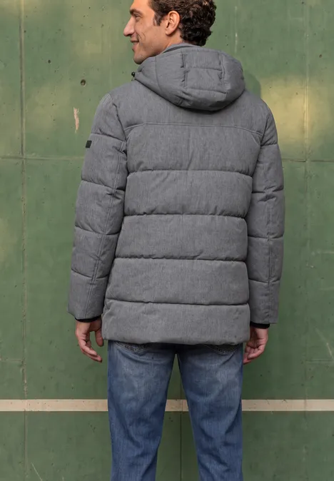 Mann von hinten in grauer, wattierter Jacke mit Kapuze und blauen Jeans vor einer grünen Wand.