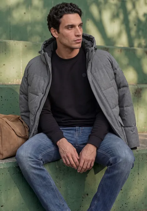 Ein Mann in grauer Steppjacke und Jeans sitzt auf einer grünen Mauer im Schatten.