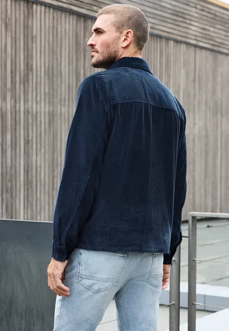 Ein Mann in dunkelblauem Cord-Hemd und hellen Jeans steht auf einer Dachterrasse vor einer Betonfassade.