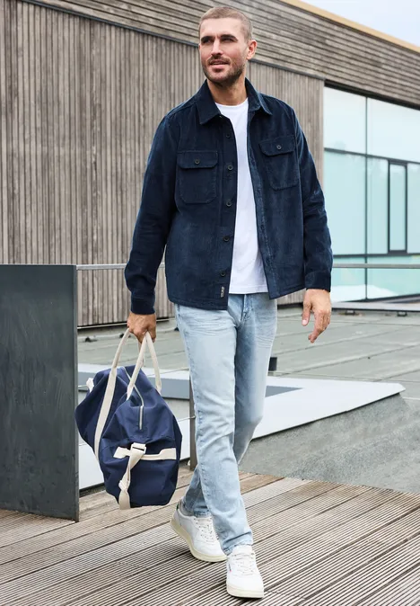 Ein Mann in Jeansjacke, weißem T-Shirt und hellen Jeans geht mit Reisetasche über eine Holzterrasse.
