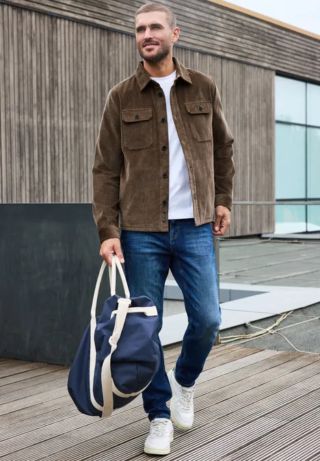 Ein Mann in brauner Jacke und Jeans steht auf einem Holzsteg und hält eine blaue Reisetasche.