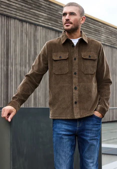 Ein Mann lehnt vor einer Holzwand, trägt eine braune Kordjacke und blaue Jeans.