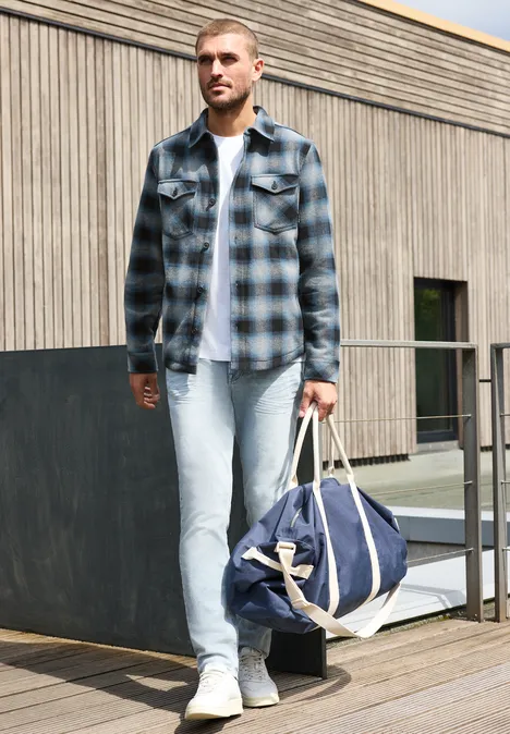 Ein Mann in kariertem Hemd und Jeans trägt eine Reisetasche auf einer Terrasse vor einem Holzhaus.