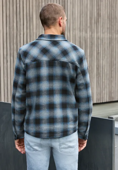 Ein Mann steht von hinten in blau-schwarz kariertem Hemd und hellen Jeans vor einer Holzfassade.