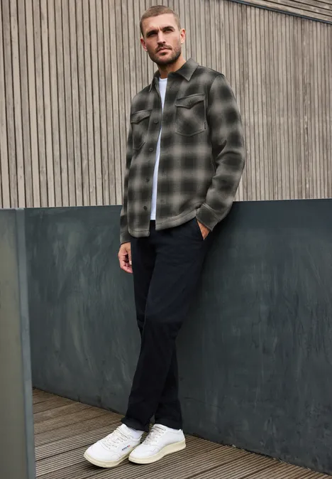 Ein Mann lehnt vor grauer Fassade, trägt kariertes Hemd, schwarze Hose und weiße Sneaker