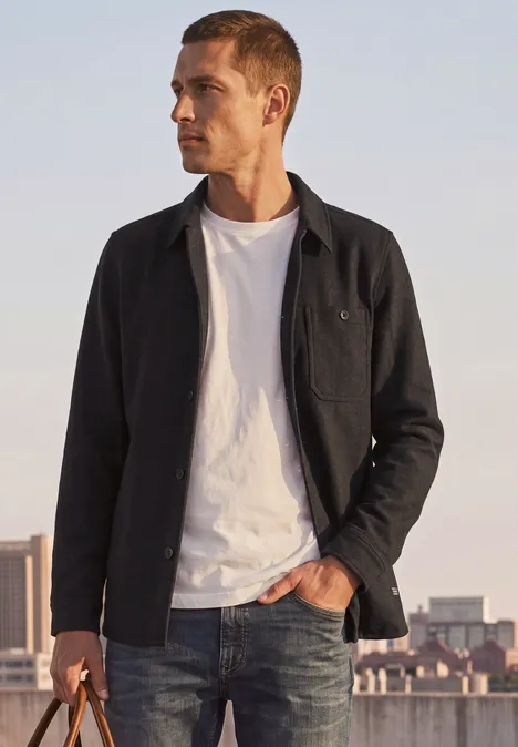 Junger Mann in Jeans, weißem T-Shirt und dunkler Jacke steht vor einer Stadt-Silhouette im Abendlicht.
