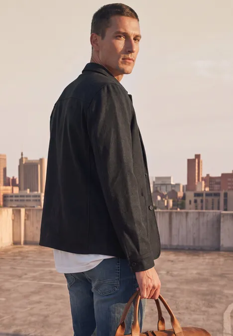 Ein Mann auf einem Dach mit Stadtpanorama im Hintergrund, trägt Jacke, Jeans und hält eine Tasche.