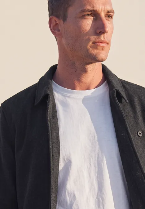 Ein nachdenklicher Mann im weißen T-Shirt und dunklem Hemd vor hellem Hintergrund, Blick seitlich.