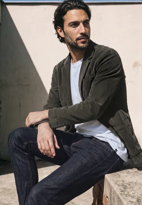 Un homme en veste de daim, T-shirt et jean est assis décontracté sur un mur en béton sous le soleil.