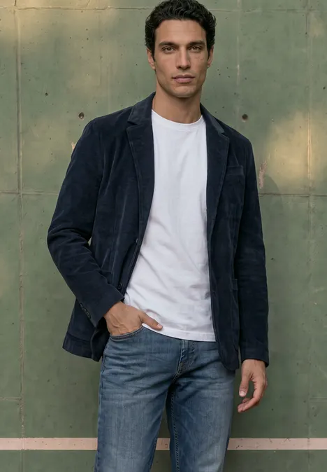 Un homme en jean, tee-shirt blanc et veste bleu foncé devant un mur vert.