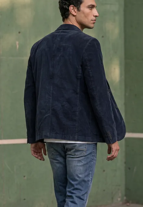 Un homme en jean et veste en velours bleu foncé se tient devant un mur vert en regardant par-dessus son épaule.