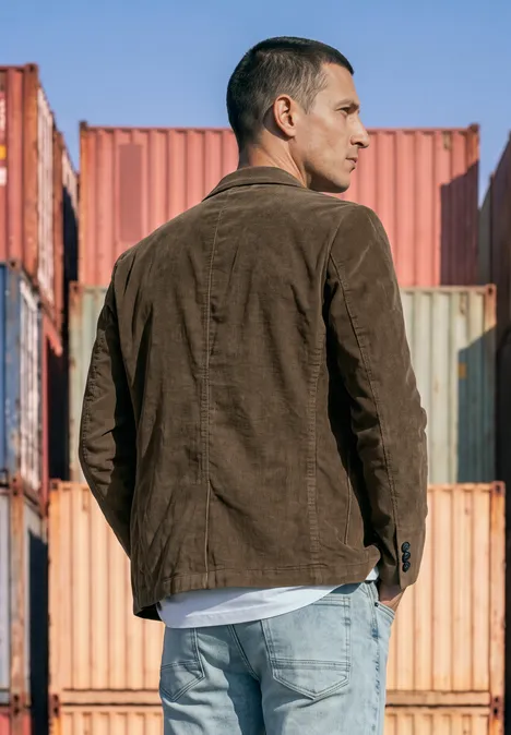 Un homme en veste marron et jean se tient de profil devant des conteneurs colorés.