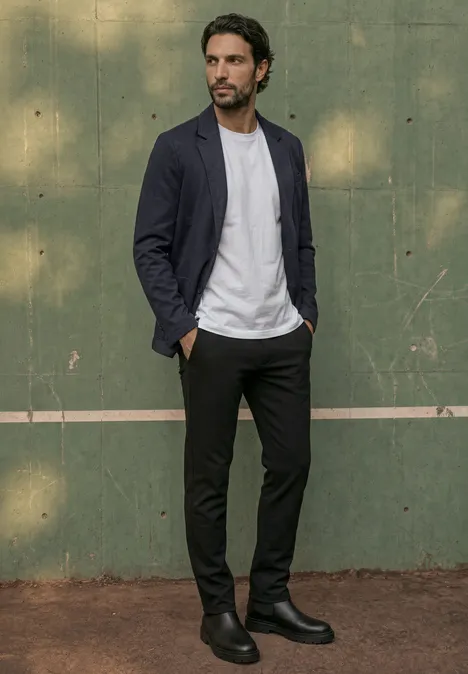 Ein Mann in legerem Outfit (Blazer, weißes T-Shirt, schwarze Hose, Boots) steht vor einer grünen Wand.